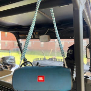 May include: A blue JBL portable speaker suspended by a white and teal braided rope strap. The speaker is cylindrical with a red JBL logo on the front. The rope is attached to the top frame of a vehicle, allowing the speaker to hang freely.