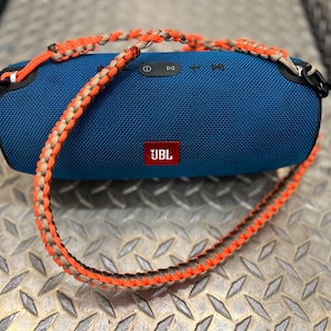 May include: A cylindrical blue JBL portable speaker featuring a red logo on the front and a black control panel on top. The speaker is attached to a braided paracord carrying strap with alternating orange and tan colours and black metal clips.
