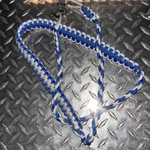 May include: A braided paracord lanyard featuring a repeating pattern of royal blue and silver-grey cord. The lanyard is constructed with a flat, wide weave section and a thinner, rounded braided section, finished with a silver-toned metal shackle clasp.