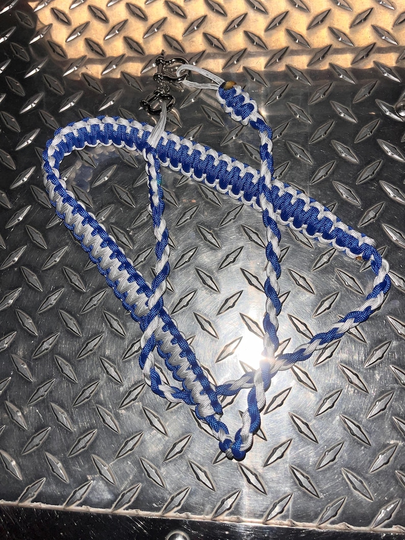 May include: A braided paracord lanyard featuring a repeating pattern of royal blue and silver-grey cord. The lanyard is constructed with a flat, wide weave section and a thinner, rounded braided section, finished with a silver-toned metal shackle clasp.