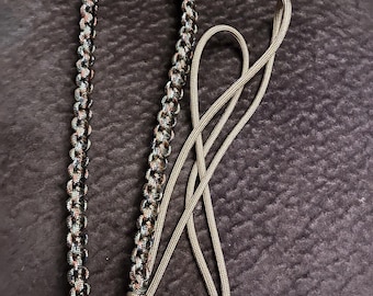 May include: A lanyard constructed from braided paracord featuring a multi-coloured pattern of black, brown, and tan tones. The braided section transitions into two smooth, solid tan cords that extend outward, forming a loop at the base of the braid.