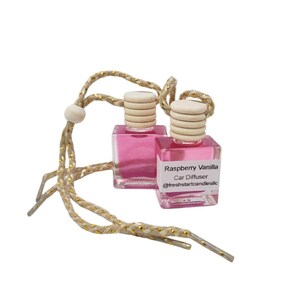 Puede incluir: Dos pequeños ambientadores cuadrados para coche llenos de líquido rosa, cada uno con una tapa de madera. Las etiquetas dicen "Raspberry Vanilla Car Diffuser" y el nombre de usuario de Instagram. Cuelgan de un cordón dorado y beige.
