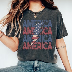 Op de afbeelding: Donkergrijs T-shirt met het woord "AMERICA" in rode en blauwe blokletters. Een bliksemflits-afbeelding met een sterren- en strepenontwerp is gecentreerd op het shirt. Het shirt wordt gecombineerd met een blauwe jeans en witte sneakers.