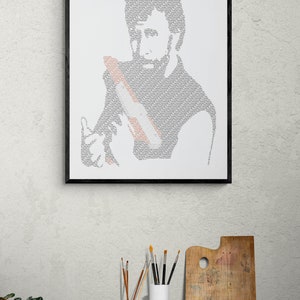 May include: A black framed print of a portrait of a person made from the word "Occasionally" repeated many times. The print is hanging on a white wall. There are paintbrushes in a white and terracotta jar on a wooden table in front of the print.