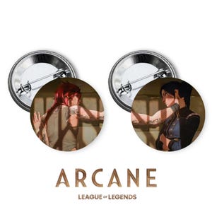 May include: Two round pinback buttons featuring characters from the animated series Arcane, based on the video game League of Legends. The left button shows a woman with red hair and tattoos, while the right button shows a woman with dark hair and a blue jacket.