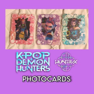 May include: Three photocards with character images in decorative frames. The frames are pink, purple, and teal, adorned with hearts and floral designs. The text "K-POP DEMON HUNTERS" and "PHOTOCARDS" are displayed below the cards, along with the "HUNTRAX" logo.