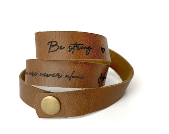 Puede incluir: Una pulsera envolvente de cuero marrón con las palabras "Be strong" y "You are never alone" grabadas en negro. La pulsera tiene un cierre de botón a presión con un botón dorado.