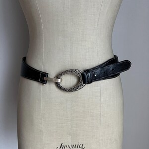 Puede incluir: Cintur&oacute;n de cuero negro con una hebilla ovalada plateada con un dise&ntilde;o ornamentado. El cintur&oacute;n tiene costuras blancas a lo largo de los bordes y se muestra en un maniqu&iacute;. La hebilla mide aproximadamente 7.6 cm de largo.
