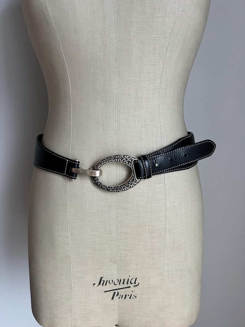 Puede incluir: Cintur&oacute;n de cuero negro con una hebilla ovalada plateada con un dise&ntilde;o ornamentado. El cintur&oacute;n tiene costuras blancas a lo largo de los bordes y se muestra en un maniqu&iacute;. La hebilla mide aproximadamente 7.6 cm de largo.