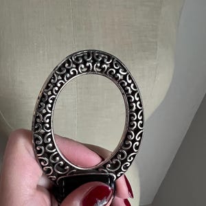 Puede incluir: Un cintur&oacute;n de cuero negro con una hebilla ovalada plateada. La hebilla tiene un dise&ntilde;o calado con un patr&oacute;n de remolino. El cintur&oacute;n est&aacute; sujeto por una mano con esmalte de u&ntilde;as rojo.