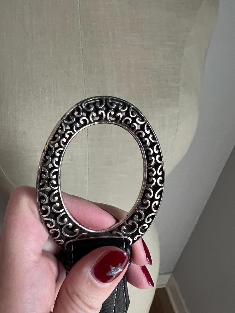 Puede incluir: Un cintur&oacute;n de cuero negro con una hebilla ovalada plateada. La hebilla tiene un dise&ntilde;o calado con un patr&oacute;n de remolino. El cintur&oacute;n est&aacute; sujeto por una mano con esmalte de u&ntilde;as rojo.