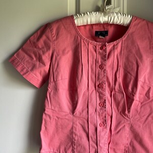 May include: A short-sleeved, button-down shirt in a solid coral pink color. The shirt features a round neckline, vertical pleats running down the front placket, and five matching pink buttons. A dark label is visible inside the collar area.