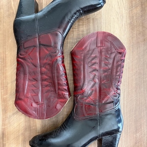 May include: A pair of two-toned cowboy boots featuring a glossy black finish on the foot and heel, transitioning into a deep red shaft. The upper shaft is decorated with intricate tonal stitching patterns and includes pull tabs at the top edges.