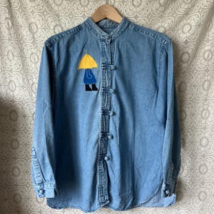 May include: Light blue denim shirt with a mandarin collar and long sleeves. The shirt features a yellow umbrella and a person in blue rain gear applique on the front. Fastened with small, round buttons.