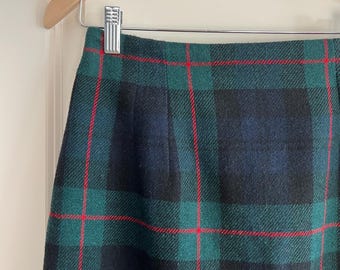 May include: A close-up of a green, navy, and red plaid skirt hanging on a wooden hanger. The skirt has a classic tartan pattern with intersecting lines of red against a backdrop of green and navy blue.