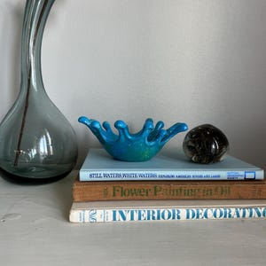 May include: A blue and green decorative bowl with a unique shape sits atop a stack of books. A tall, gray glass vase with a flower and a decorative glass sphere are also present. The books include titles like "Still Waters, White Waters" and "Flower Painting in Oil."