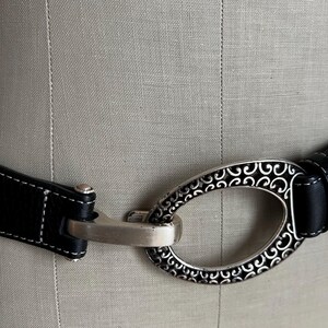 Puede incluir: Cintur&oacute;n de cuero negro con una hebilla ovalada plateada con un dise&ntilde;o ornamental de volutas. El cintur&oacute;n tiene costuras blancas a lo largo de los bordes y un cierre plateado. La hebilla mide unos 7,6 cm de largo.