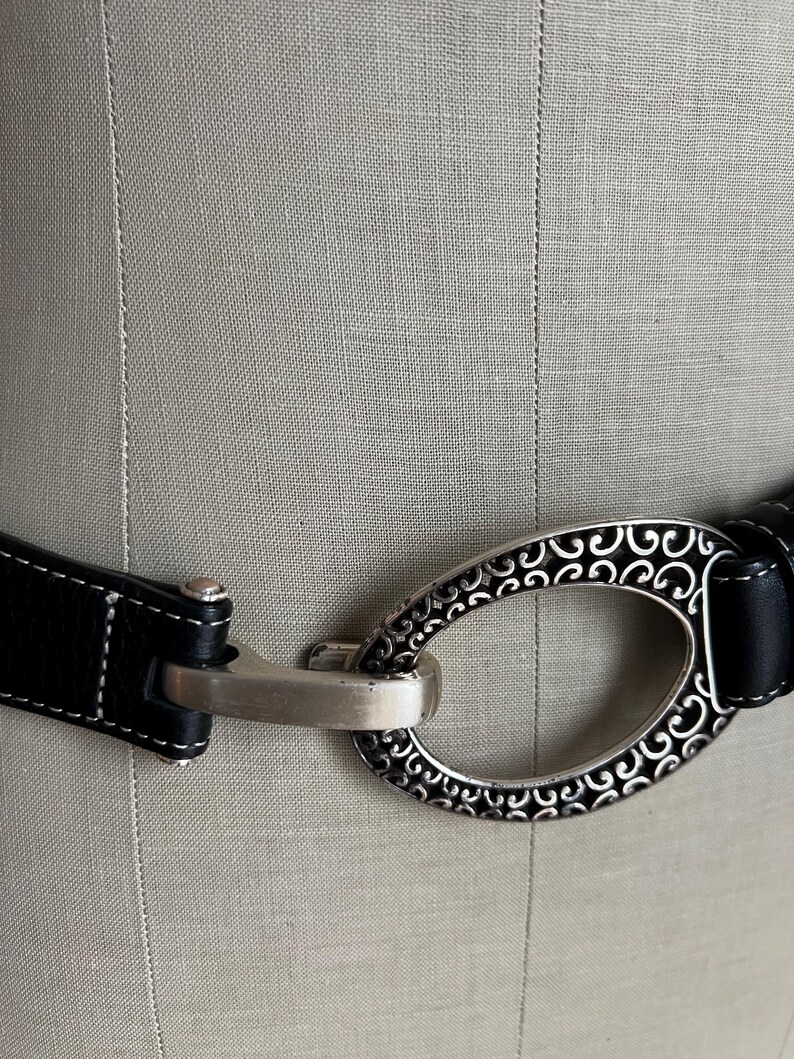 Puede incluir: Cintur&oacute;n de cuero negro con una hebilla ovalada plateada con un dise&ntilde;o ornamental de volutas. El cintur&oacute;n tiene costuras blancas a lo largo de los bordes y un cierre plateado. La hebilla mide unos 7,6 cm de largo.