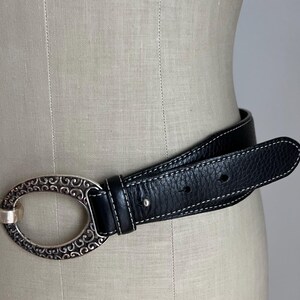 Puede incluir: Cintur&oacute;n de cuero negro con una hebilla ovalada plateada con un dise&ntilde;o de volutas ornamentadas. El cintur&oacute;n tiene costuras blancas a lo largo de los bordes. El cintur&oacute;n se muestra en un maniqu&iacute;.