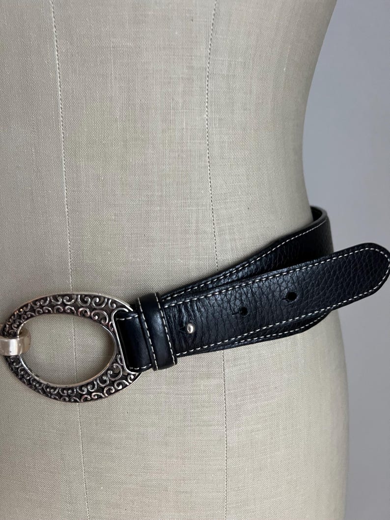 Puede incluir: Cintur&oacute;n de cuero negro con una hebilla ovalada plateada con un dise&ntilde;o de volutas ornamentadas. El cintur&oacute;n tiene costuras blancas a lo largo de los bordes. El cintur&oacute;n se muestra en un maniqu&iacute;.