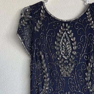 May include: A navy blue dress with silver beading and sequins. The dress features a round neckline and short sleeves. The intricate beadwork creates a floral and swirling pattern across the bodice and sleeves. The dress is hanging on a white hanger.