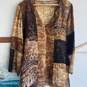 May include: A long-sleeved blouse with a V-neck and a patchwork design in shades of brown, tan, and black. The blouse is hanging on a wooden hanger. The fabric has various patterns and textures.