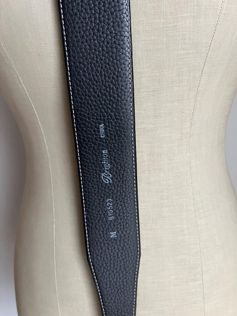 Puede incluir: Cintur&oacute;n de cuero negro con costuras blancas. El cintur&oacute;n tiene una superficie texturizada y la marca "Brighton" es visible. El cintur&oacute;n est&aacute; marcado con "M", "B10523" y "CHINA".