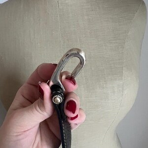 Puede incluir: Una hebilla de cintur&oacute;n plateada con un dise&ntilde;o curvo, sujeta a un cintur&oacute;n de cuero negro. El cintur&oacute;n tiene costuras blancas a lo largo de los bordes y un remache dorado. La hebilla se sostiene frente a un maniqu&iacute; beige.