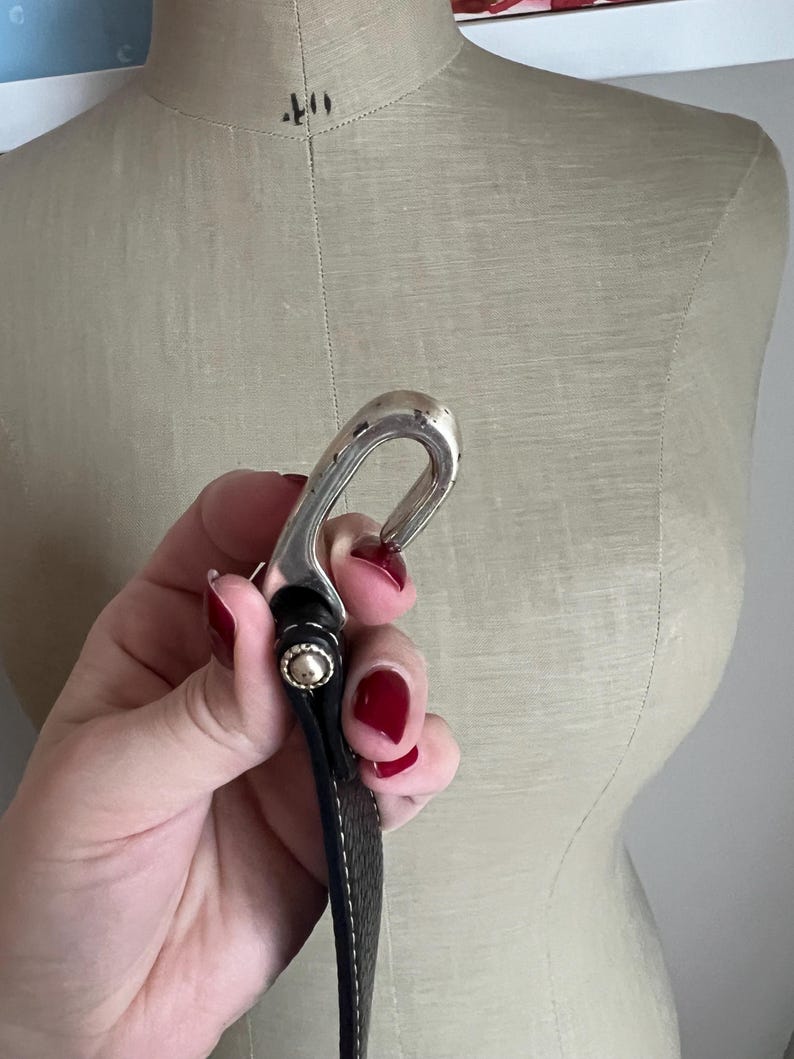 Puede incluir: Una hebilla de cintur&oacute;n plateada con un dise&ntilde;o curvo, sujeta a un cintur&oacute;n de cuero negro. El cintur&oacute;n tiene costuras blancas a lo largo de los bordes y un remache dorado. La hebilla se sostiene frente a un maniqu&iacute; beige.