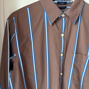 May include: A brown button-down shirt featuring vertical stripes in white and bright blue. The shirt has a pointed collar, a front chest pocket, and light-colored buttons. A dark blue label inside the collar displays the word CLASSIC.