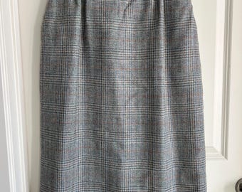 May include: A knee-length skirt featuring a grey, blue, and rust-colored plaid pattern. The skirt has a high-waisted design with subtle pleats at the waistband and hangs straight down, suspended from a black plastic clothing hanger.