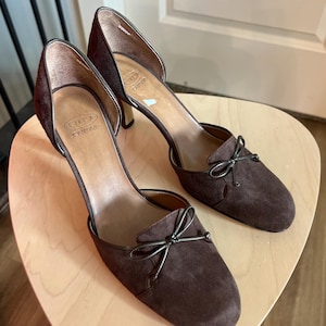 May include: A pair of brown suede heels with a bow detail on the toe. The heels have a high heel and a cutout design on the side. The brand name "CIRCA JOAN DAVID" is visible on the insole.