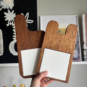 May include: Two wooden serving boards with a handle-like top and a square white tile insert. The boards are a warm brown color and are being held up in front of a wall with artwork. The boards are likely for serving appetizers or small dishes.