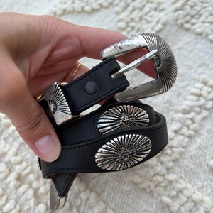 May include: A black leather belt featuring silver-toned metal hardware, including a sunburst-patterned buckle and matching oval-shaped concho accents along the strap. The belt has a smooth finish and a classic prong-style closure mechanism.