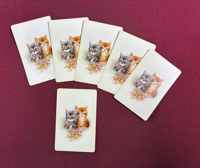 May include: A collection of vintage playing cards featuring illustrations of two kittens. One kitten is gray and the other is orange, with a floral bouquet at the bottom. The cards are arranged on a burgundy surface.