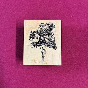 May include: A rectangular wooden rubber stamp featuring a black ink illustration of a winged fairy figure. The fairy is depicted with detailed wings and a flowing skirt, with small dots trailing from the bottom, centered on a light wood block.