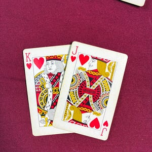 May include: Two playing cards featuring the King and Jack of Hearts. The King card shows a regal figure in red, gold, and white, while the Jack card displays a similar design. Both cards have a white background and are set against a burgundy surface.