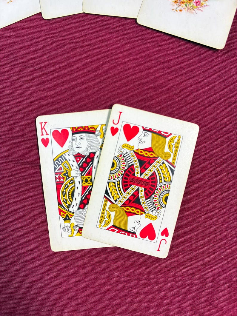 May include: Two playing cards featuring the King and Jack of Hearts. The King card shows a regal figure in red, gold, and white, while the Jack card displays a similar design. Both cards have a white background and are set against a burgundy surface.