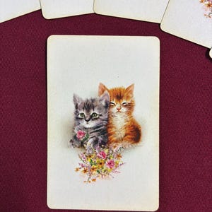 May include: A vintage playing card featuring two kittens, one gray and one orange, nestled together. They are surrounded by a bouquet of pink and yellow flowers. Several other cards with floral designs are visible.