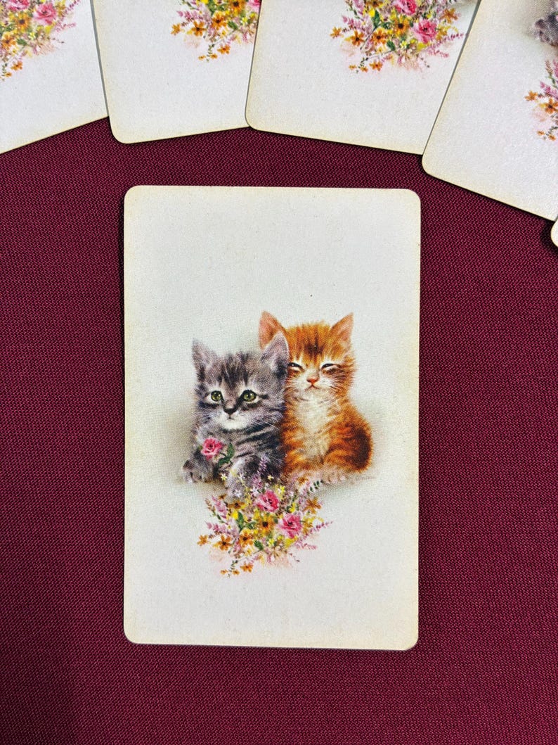 May include: A vintage playing card featuring two kittens, one gray and one orange, nestled together. They are surrounded by a bouquet of pink and yellow flowers. Several other cards with floral designs are visible.