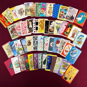 May include: A collection of vintage playing cards, each with unique illustrations. The cards feature a variety of images, including animals, flowers, portraits, and abstract designs. The cards are arranged on a burgundy surface.