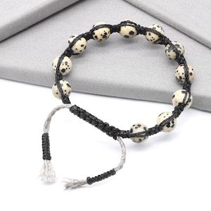 May include: A handmade bracelet featuring cream-colored, speckled beads strung on black cord. The bracelet has an adjustable closure with gray and black woven cords. The beads are approximately 0.5 inches in diameter.