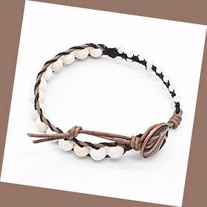 May include: A wrap bracelet featuring cream-colored beads, black and brown braided cord, and a brown leather strap. The bracelet has a copper-colored clasp with a decorative design.