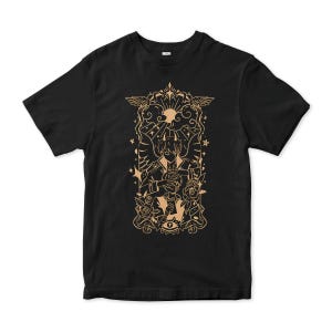 Puede incluir: Camiseta negra con un estampado gráfico dorado de una persona con alas y un halo, rodeada de flores y estrellas. El gráfico tiene un tema místico y celestial.