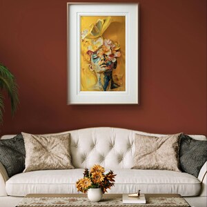 Op de afbeelding: Een ingelijst schilderij van een vrouwen gezicht met gele bloemen die haar ogen bedekken. De achtergrond is geel en oranje.