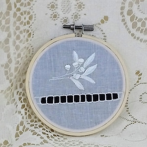 May include: A white embroidery hoop with a blue fabric background. The embroidery features a white floral design with a black ribbon.