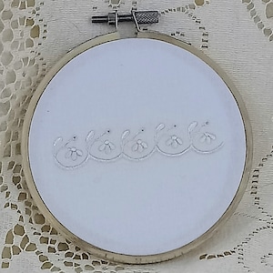 May include: White embroidery hoop with a simple white floral design stitched on the fabric.