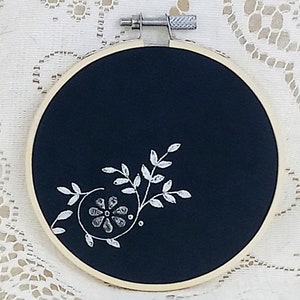 May include: A black embroidery hoop with a white floral design. The design features a stylised flower with leaves.