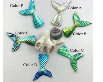 以下が含まれることがあります： 6つの異なる色の虹色のマーメイドテールのチャーム。A - 白、B - 緑、C - 緑、D - 青、E - 黄、F - 青。チャームは白い背景に円形に配置されています。