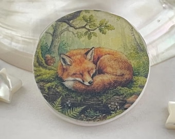 以下が含まれることがあります： 森の中で眠るキツネを描いた丸い装飾品。オレンジ色の毛並みのキツネが、緑色の苔とシダの上に丸まっています。背景には木とリスが描かれています。このアイテムには白い縁があります。